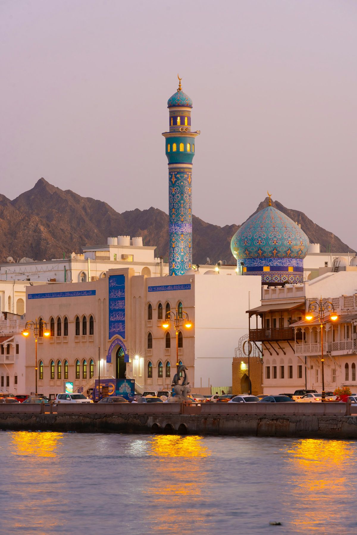 A blue-tiled mosque dome and minaret on the Muttrah waterfront in Muscat, with mountains behind